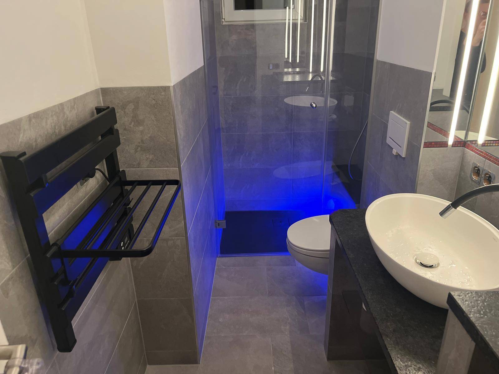 Modernes Badezimmer mit begehbarer Dusche, beheiztem Handtuchhalter und blauer Akzentbeleuchtung.