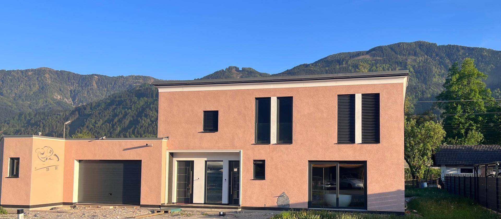 Modernes Einfamilienhaus mit rosafarbener Fassade und dunklen Fensterläden vor grüner Bergkulisse.