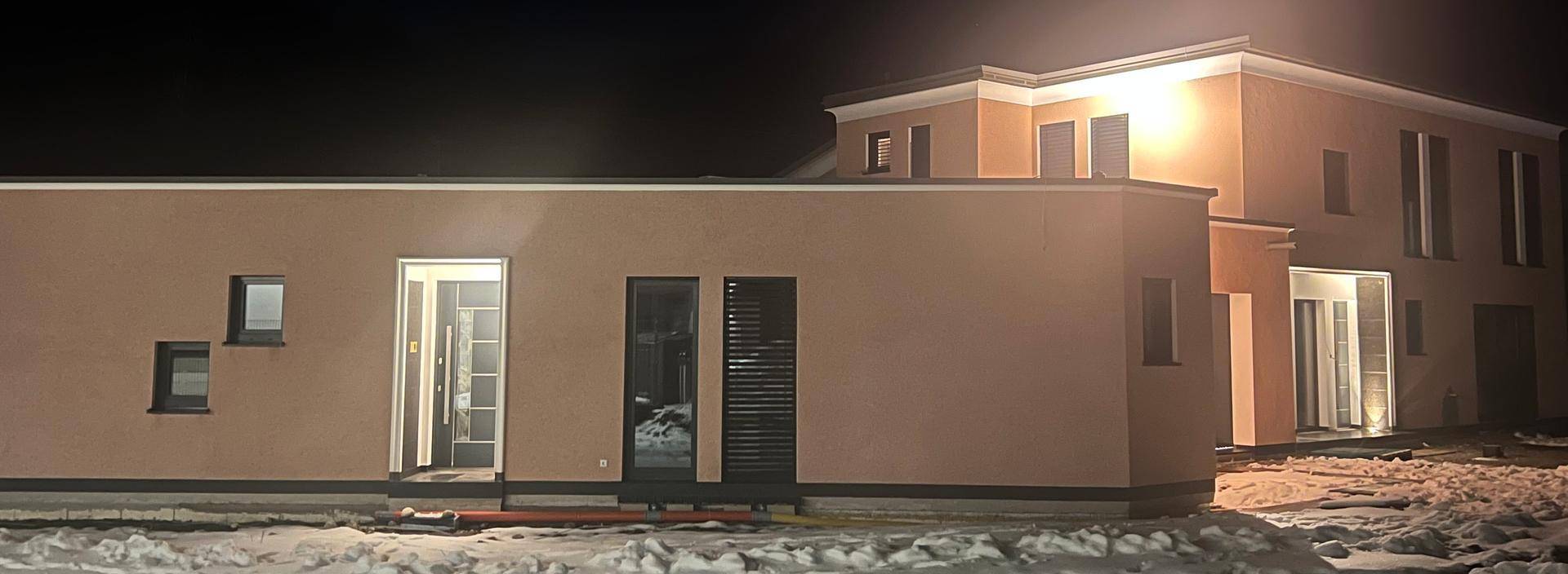 Modernes Haus mit rosafarbener Fassade und beleuchtetem Eingangsbereich bei Nacht.