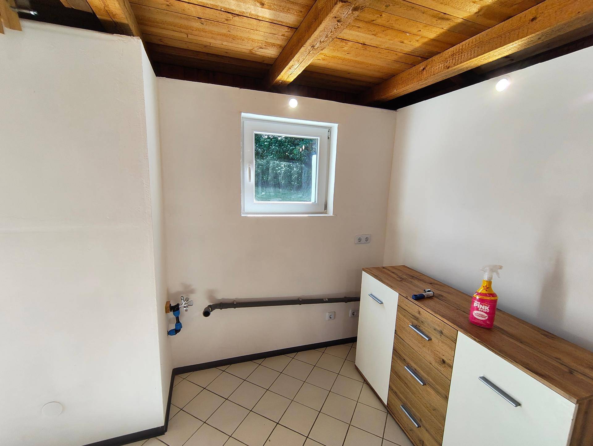 Innenansicht eines funktionalen Raumes mit Fenster, Fliesenboden und sichtbaren Wasseranschlüssen.