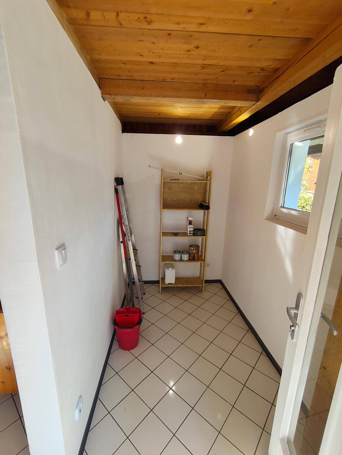 Innenraum eines Abstellraums mit Fliesenboden, Holzbalkendecke und einem kleinen Fenster.