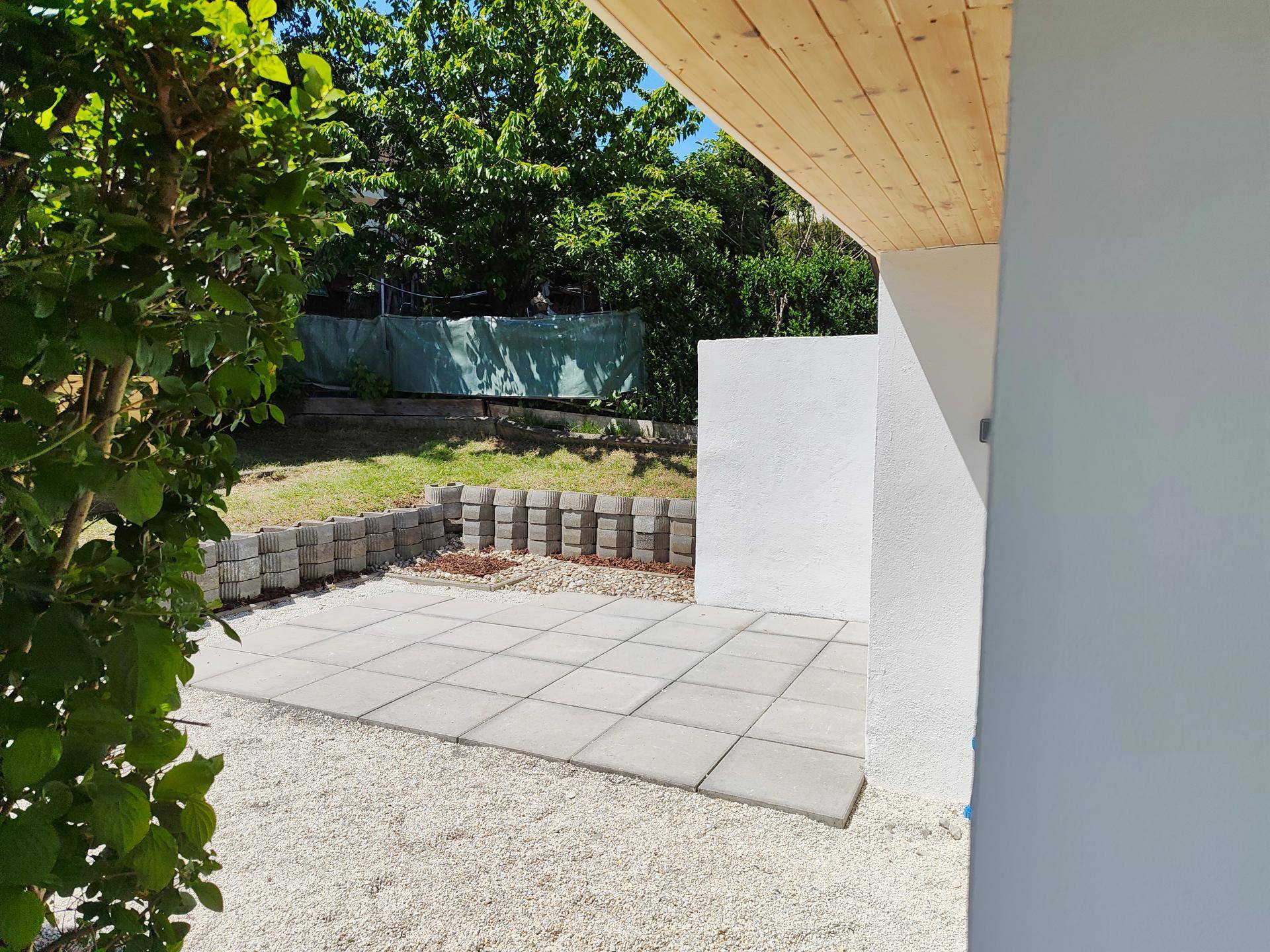 Blick von einem überdachten Bereich auf eine gepflasterte Fläche und den Garten mit grüner Bepflanzung.