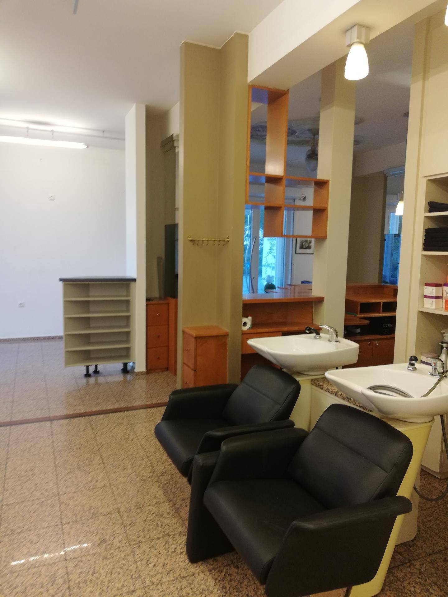 Friseursalon-Bereich mit zwei Waschbecken, schwarzen Stühlen und Regalen, verbunden mit einem weiteren Raum.