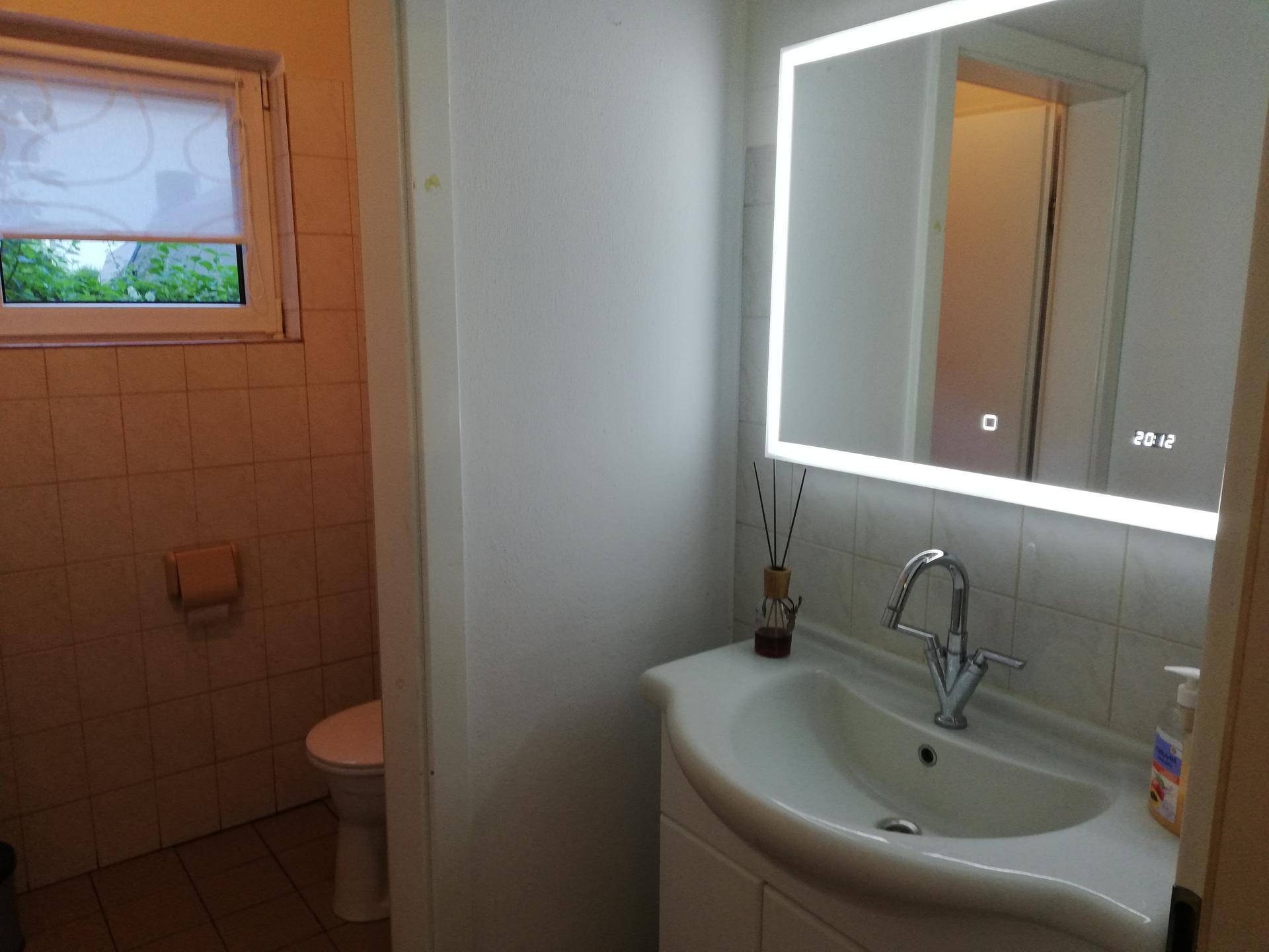 Modernes Badezimmer mit beleuchtetem Spiegel, Waschbecken und Fenster, gefliest in hellen Tönen.