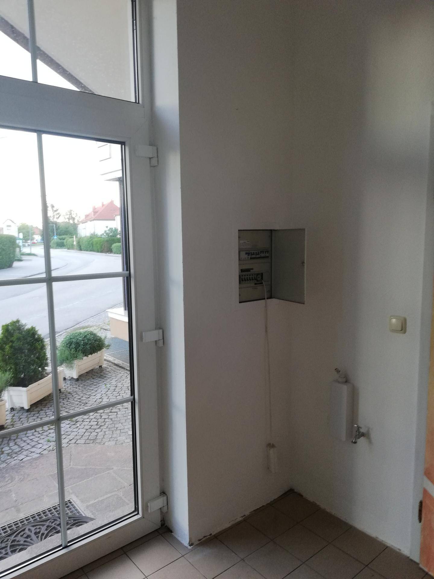 Eingangsbereich mit großer Glastür, Blick nach draußen und einem Sicherungskasten an der Wand.