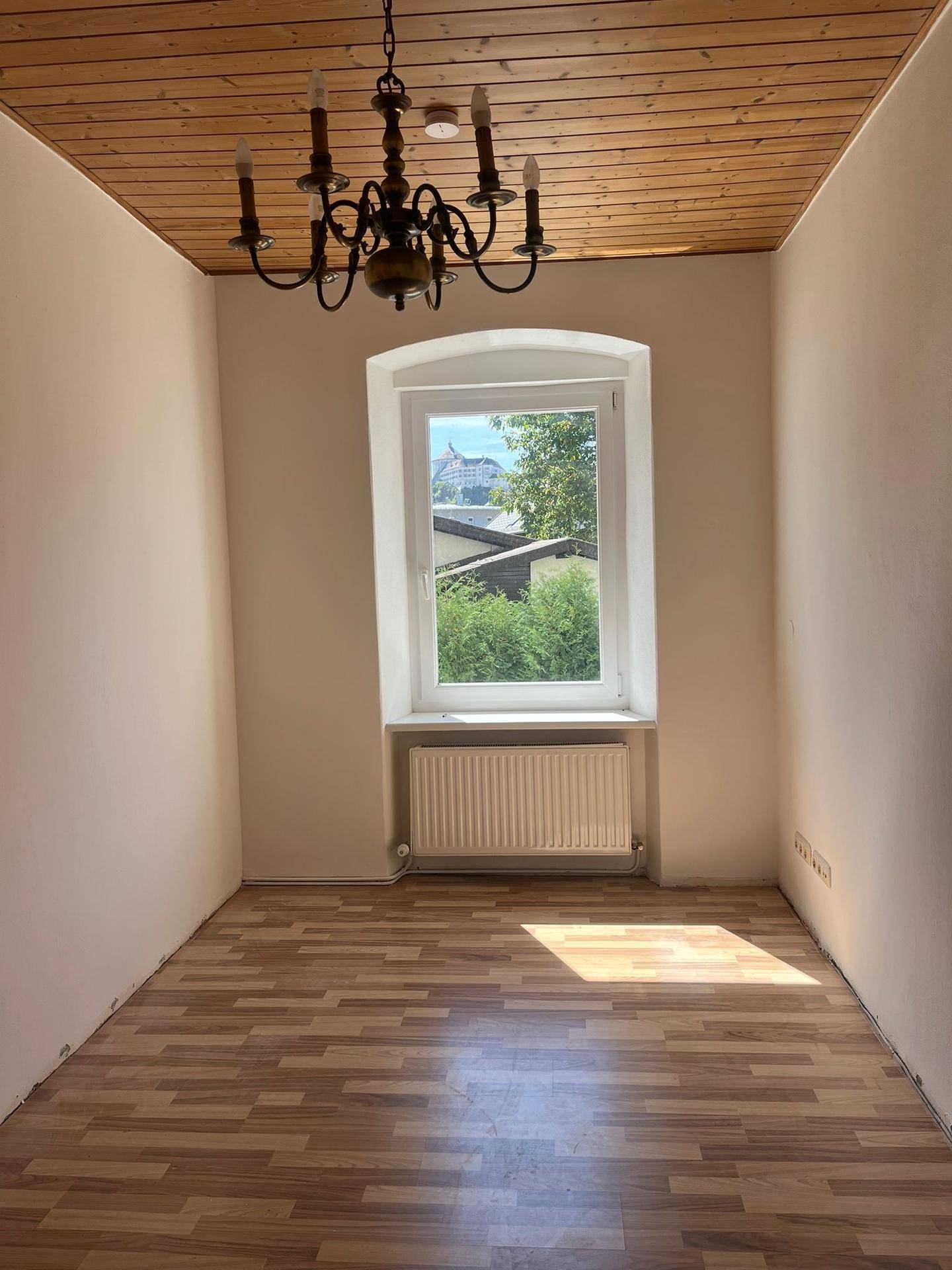 Heller leerer Raum mit Holzdecke, Laminatboden und Fenster mit Ausblick ins Grüne.