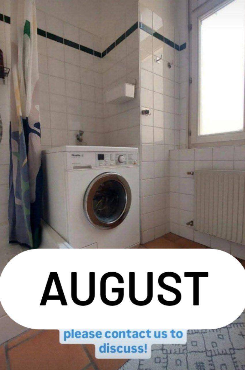 Badezimmer mit Waschmaschine und Duschvorhang, hell durch das Fenster und mit Terrakottaboden.
