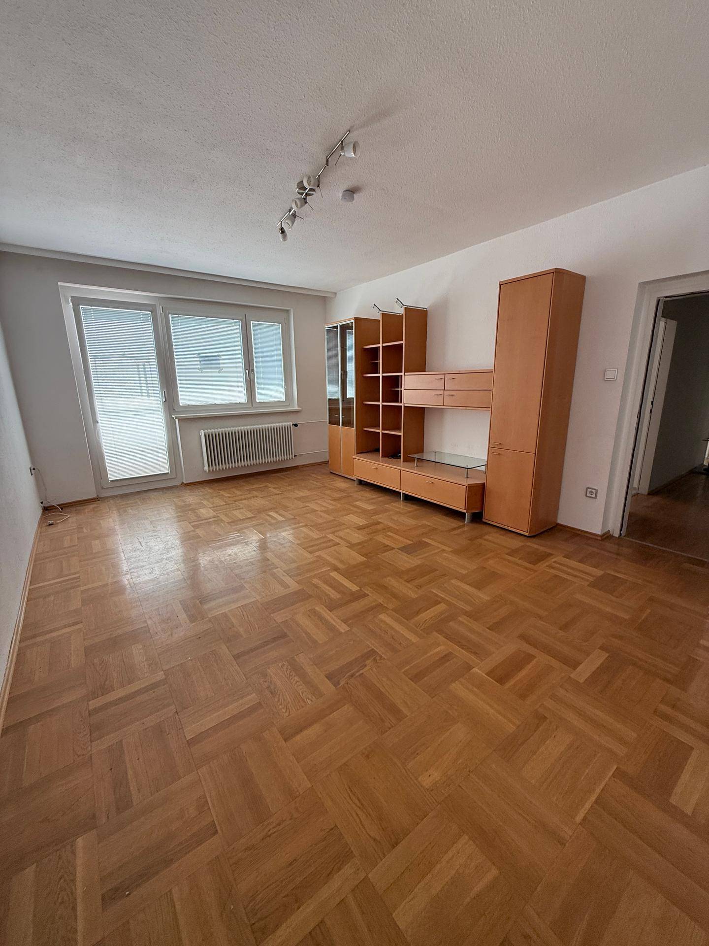 Geräumiges Wohnzimmer mit klassischem Parkettboden, großen Fenstern und Zugang zum Balkon.