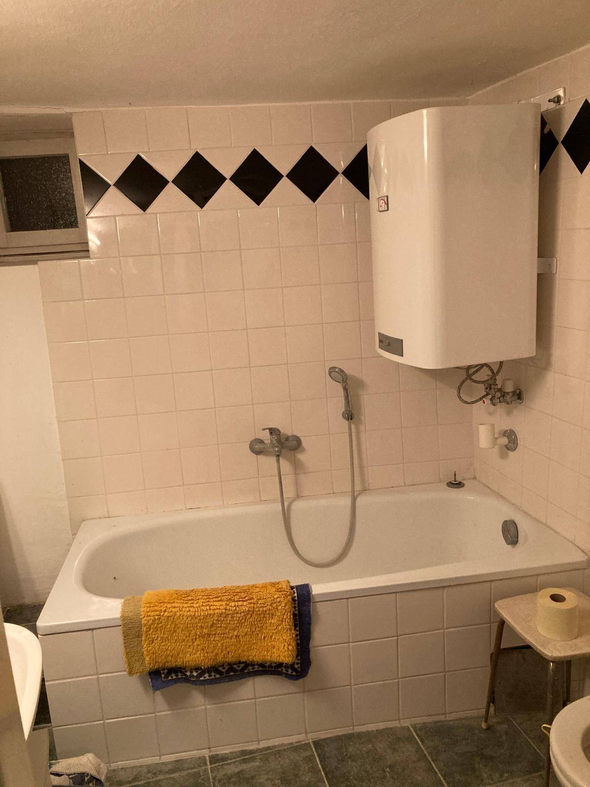 Badezimmer mit einer Badewanne, weißen Fliesen und einem Wandboiler, in einem älteren, aber gepflegten Zustand.