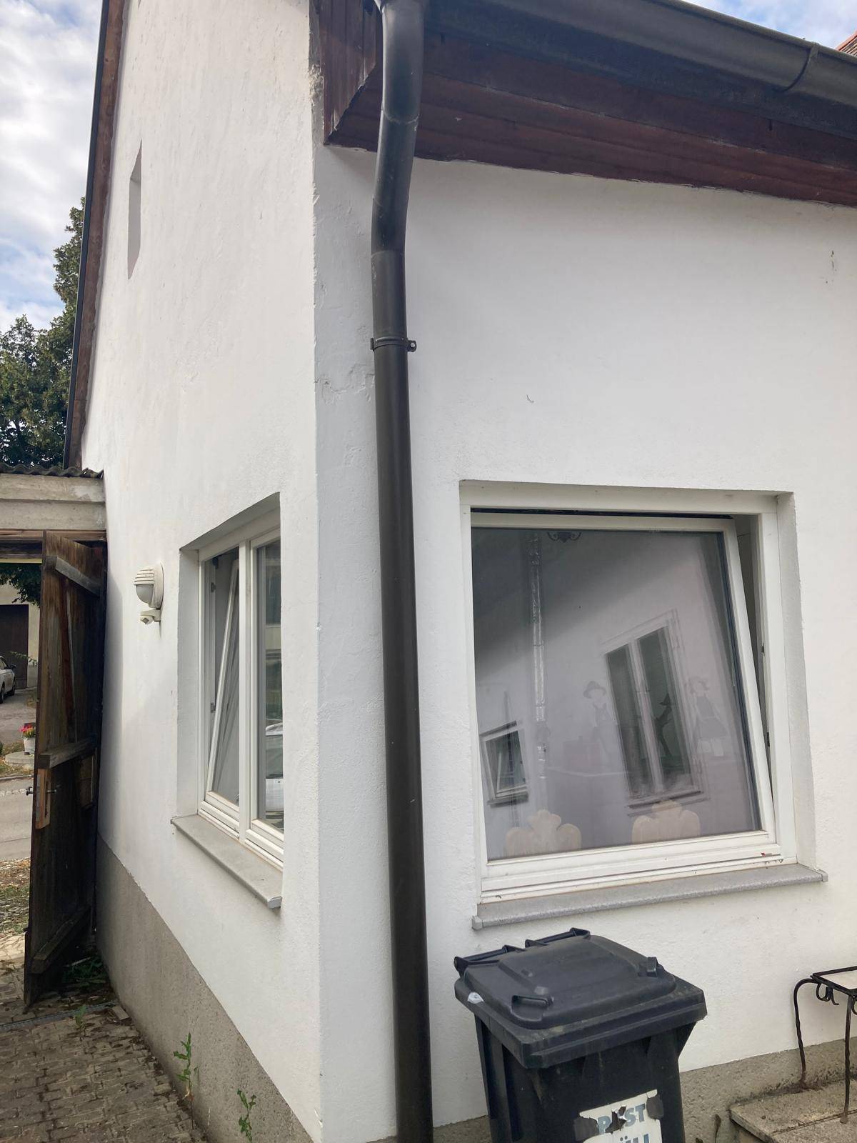 Ecke eines weißen Hauses mit Fenstern und einer Regenrinne, daneben ein Mülleimer.