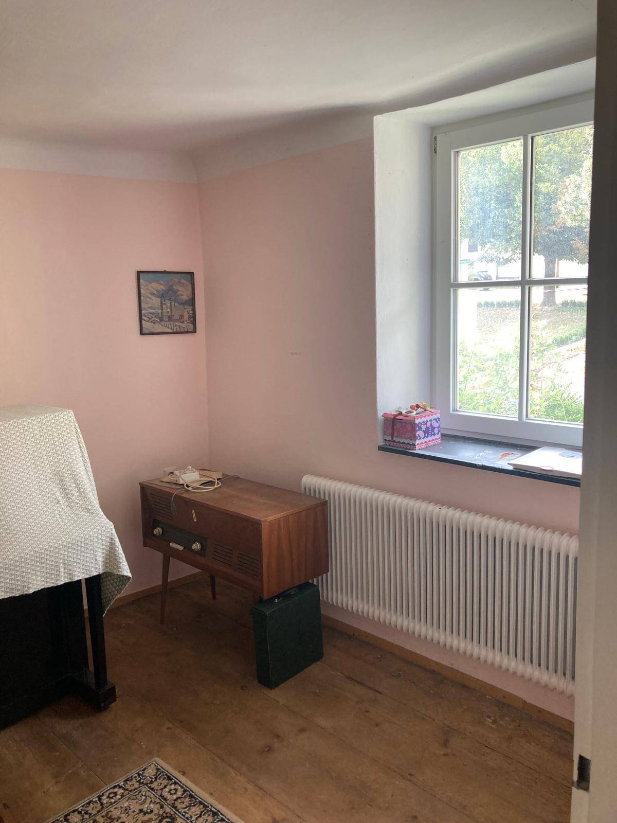 Helles Zimmer mit rosa Wänden, Holzfußboden und einem Fenster mit Blick ins Grüne.