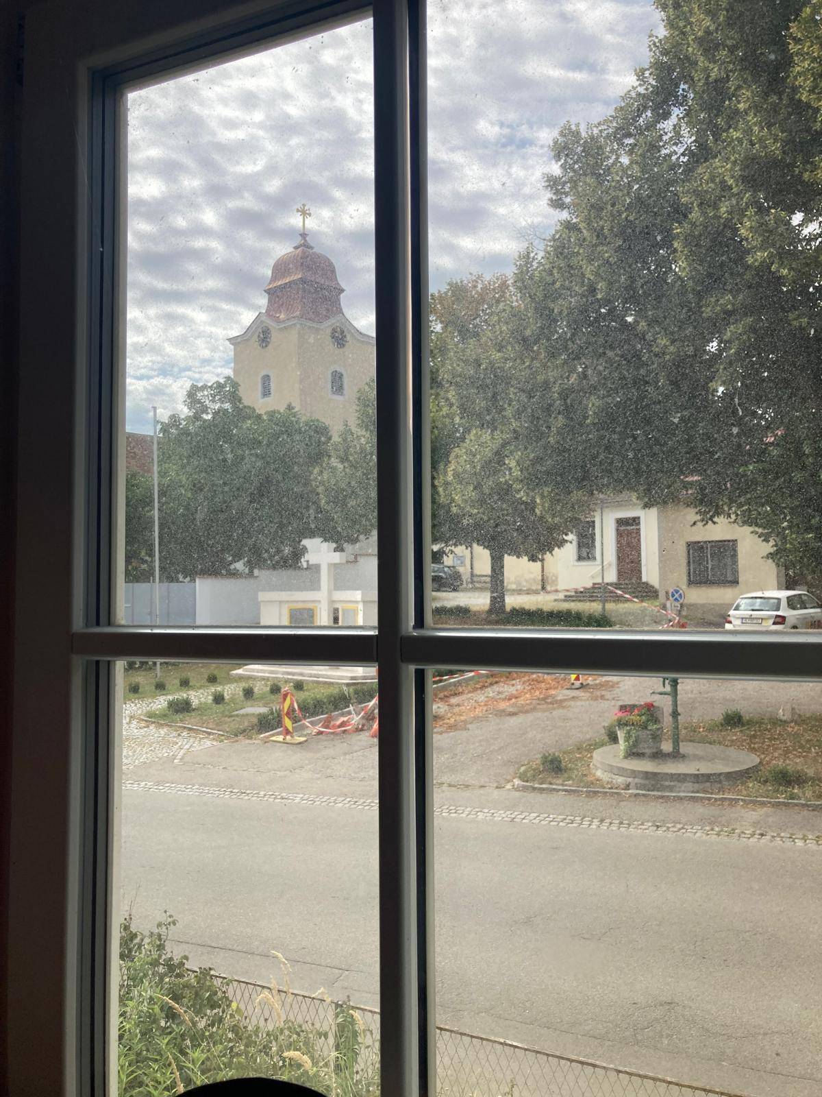 Blick aus dem Fenster auf eine ruhige Straße mit Bäumen und einem Kirchturm im Hintergrund.