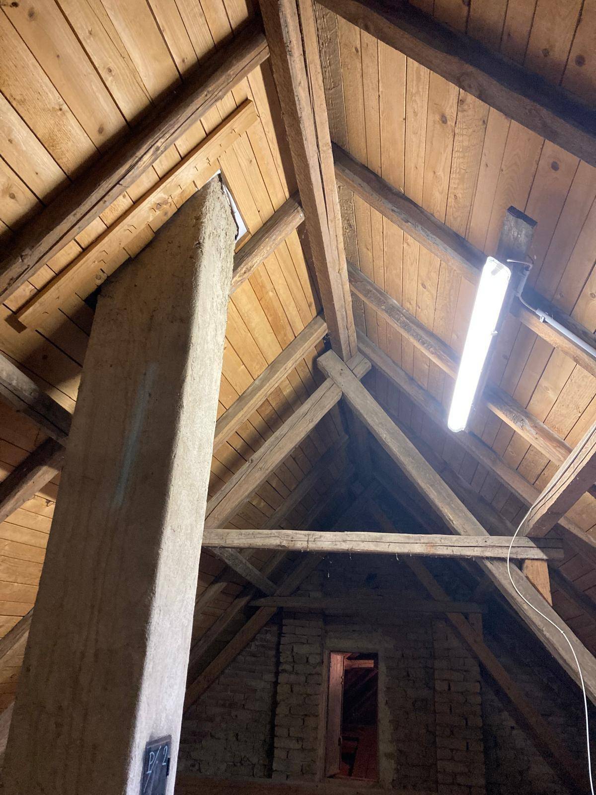 Dachboden mit freiliegenden Holzbalken und einem gemauerten Kamin, beleuchtet von einer Neonlampe.