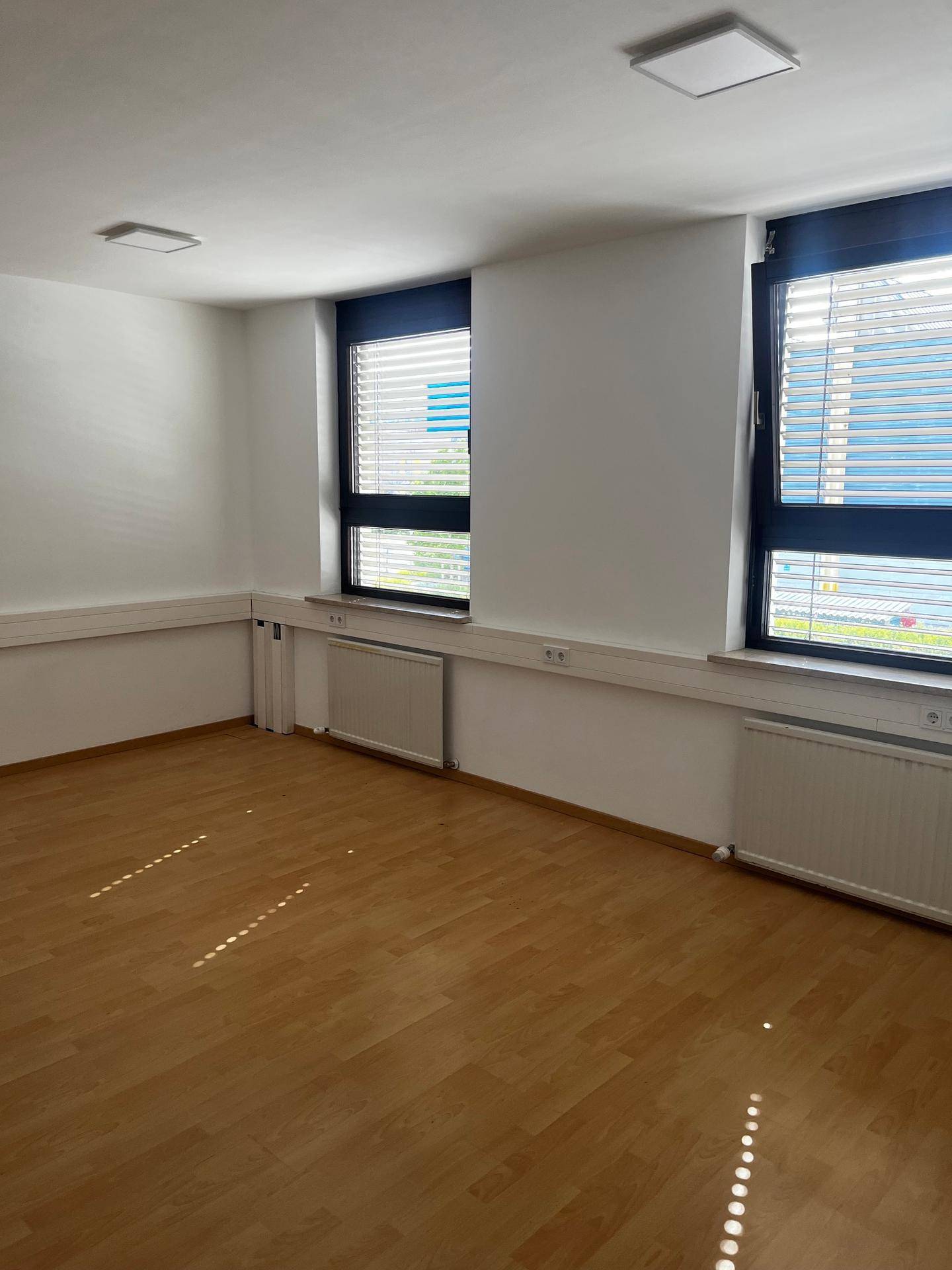 Heller Büroraum mit zwei Fenstern, Jalousien, Holzfußboden und Heizkörpern unter den Fensterbänken.
