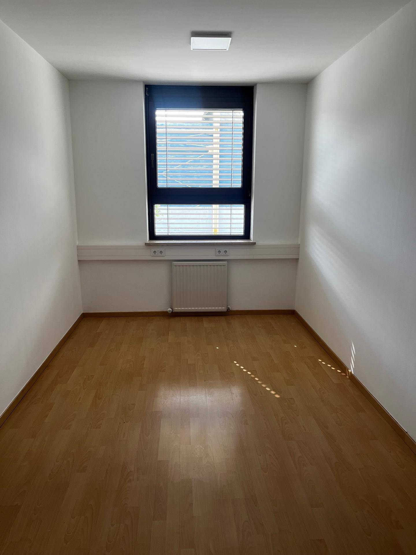 Schmaler Büroraum mit hellem Holzfußboden, einem Fenster mit Jalousien und einem Heizkörper.
