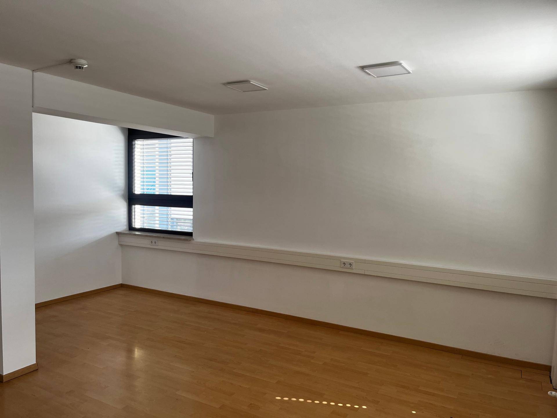 Geräumiger Büroraum mit hellem Holzfußboden, einem Fenster mit Jalousien und einem Rauchmelder an der Decke.