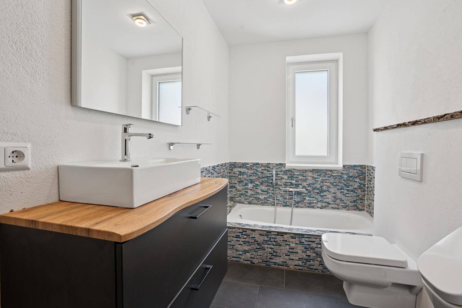 Helles Badezimmer mit Badewanne, Waschtisch und farbigen Mosaikfliesen.