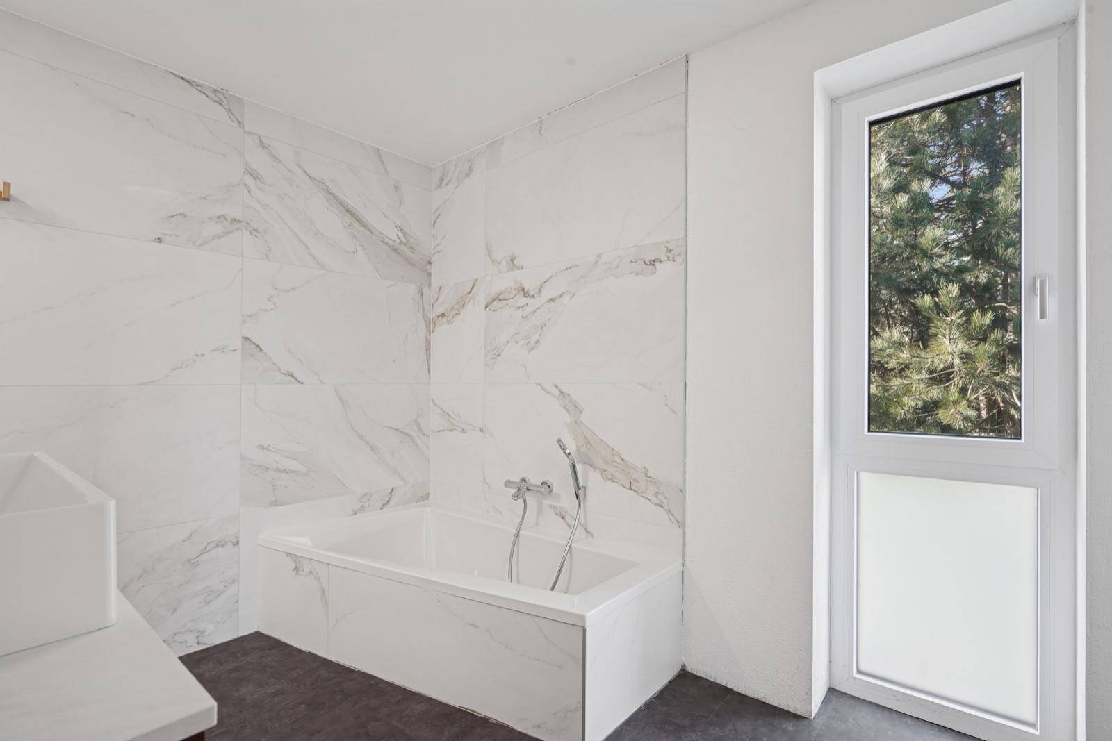Badezimmer mit Badewanne und großen Marmorfliesen, hell durch das Fenster.