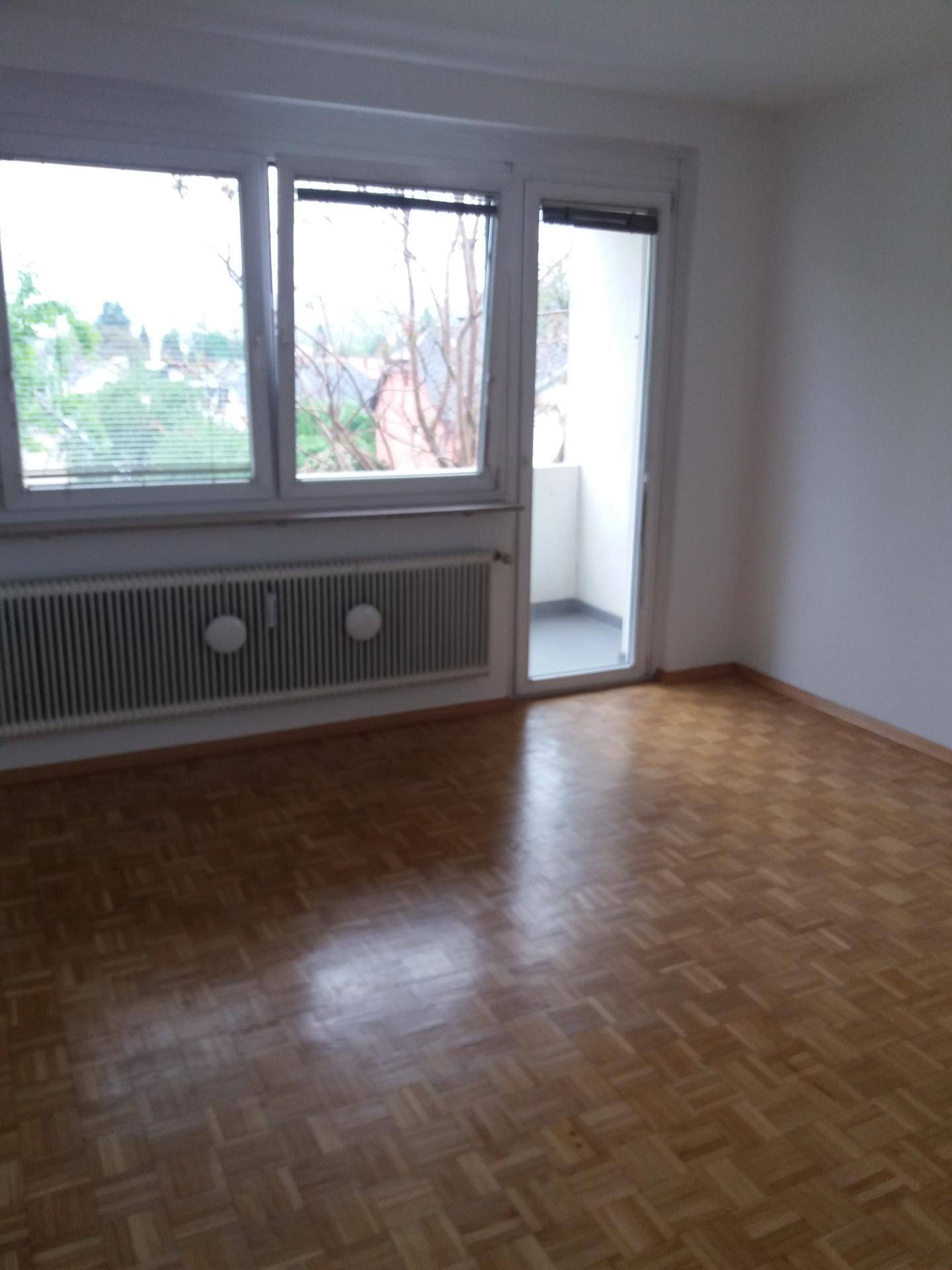 Helles Zimmer mit Parkettboden, großen Fenstern und Zugang zu einem kleinen Balkon.
