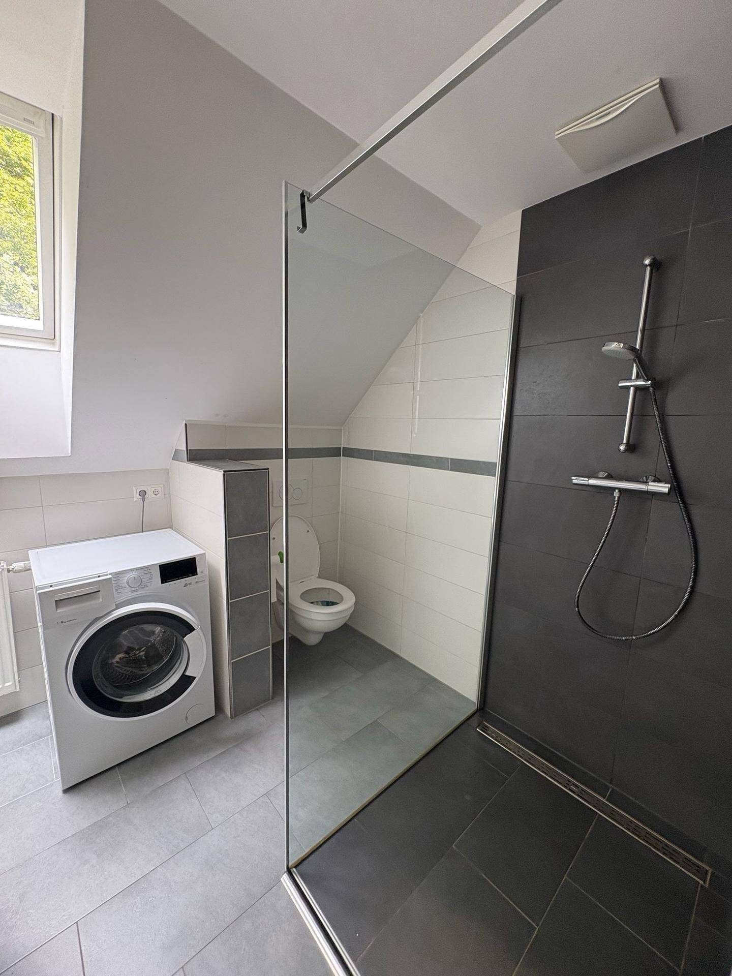 Modernes Badezimmer mit begehbarer Dusche, WC und Waschmaschine unter einer Dachschräge.
