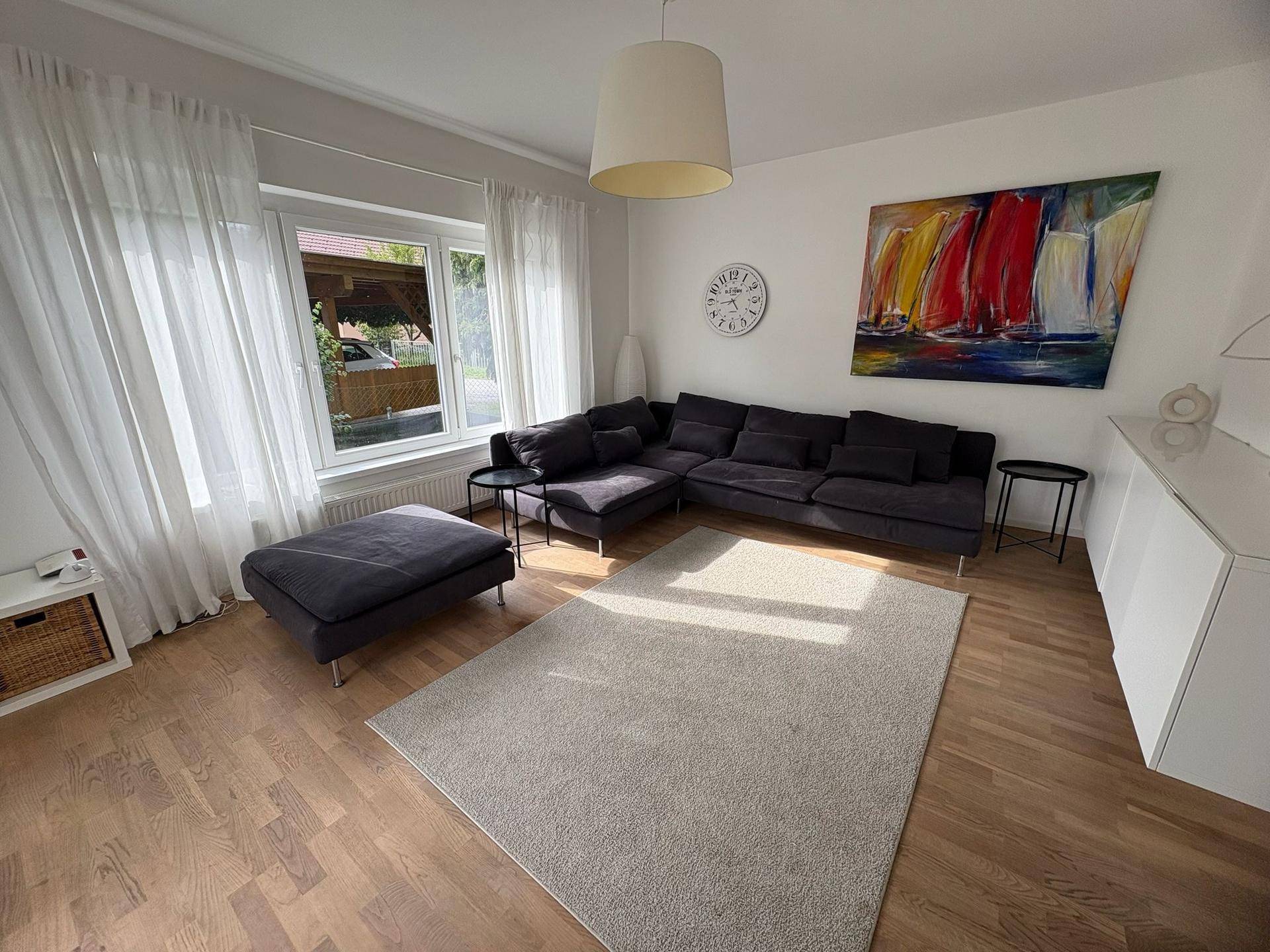 Helles Wohnzimmer mit dunklem Ecksofa, hellem Teppich und Holzboden, dekoriert mit einem farbenfrohen Bild.