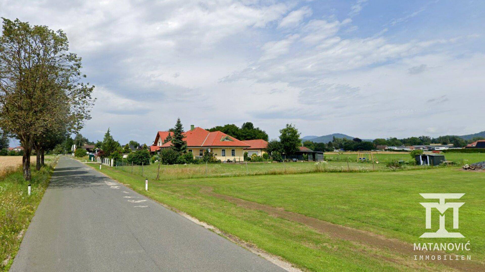 Ländliche Straße neben einer weiten Wiese mit einem Haus und Bergen im Hintergrund.