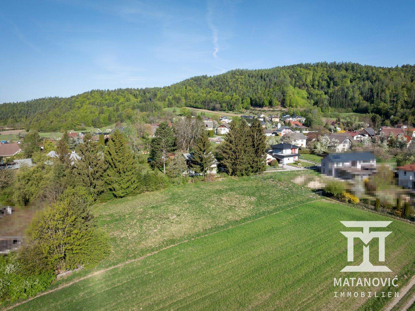 Luftaufnahme eines Feldes mit angrenzender Wohnsiedlung und bewaldeten Hügeln im Hintergrund.