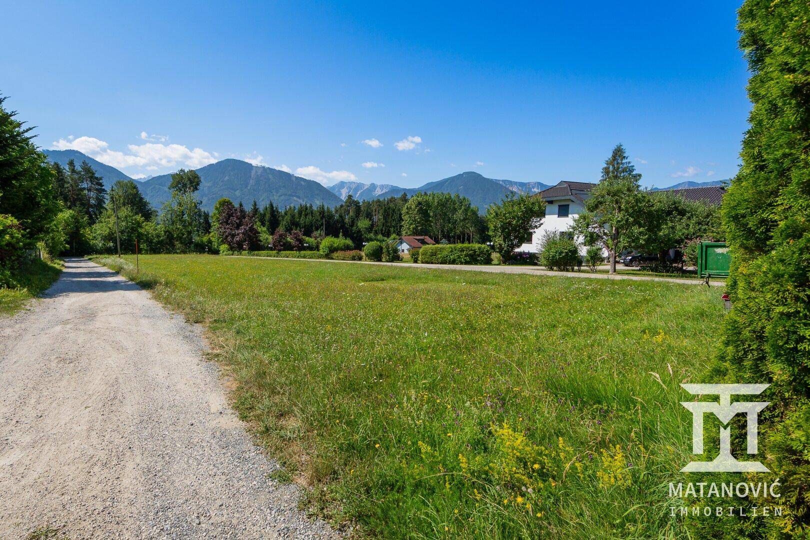 Weitläufige grüne Wiese neben einem Schotterweg, mit einem Haus und Bergen im Hintergrund.