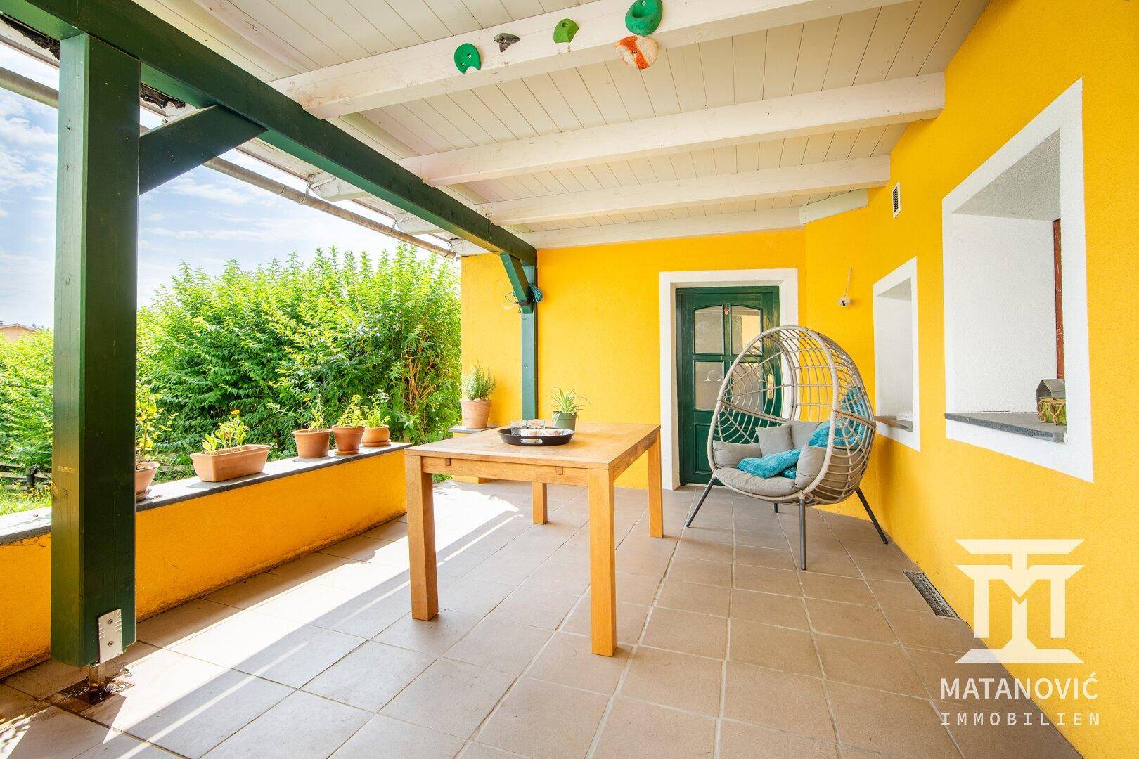 Überdachte Terrasse mit Fliesenboden, Holztisch und einem modernen Hängesessel.