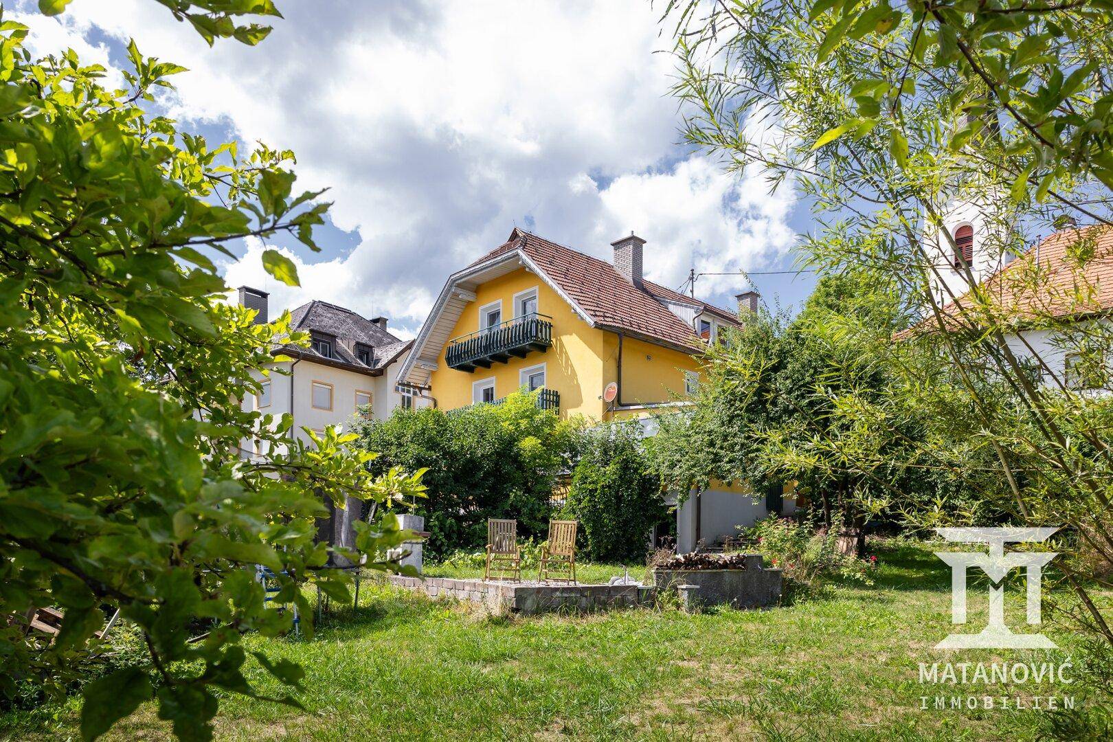 Gelbes Haus umgeben von Bäumen und Büschen, mit einem kleinen Gartenbereich und Sitzgelegenheiten.