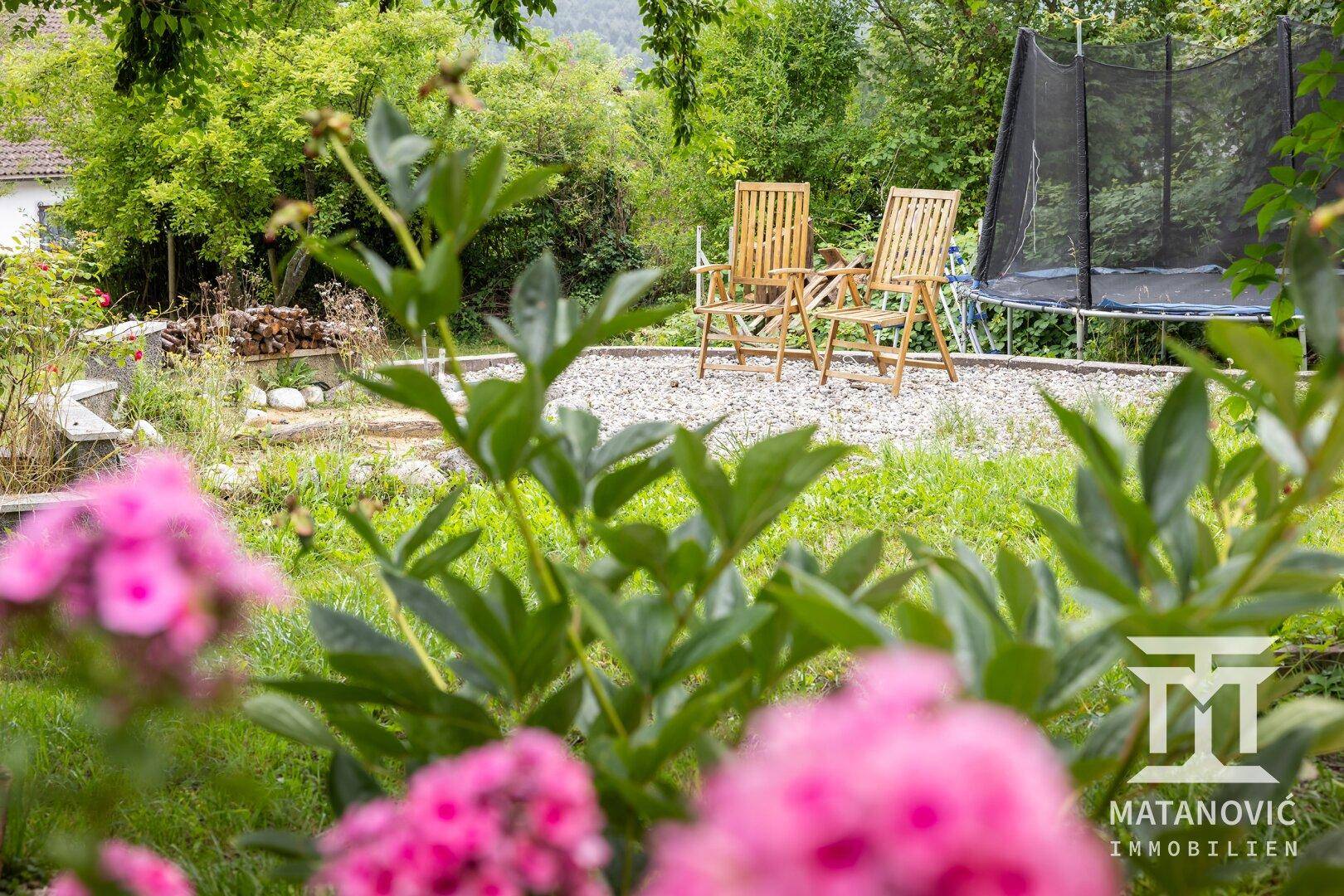 Idyllischer Gartenbereich mit blühenden Blumen, Sitzgelegenheiten und einem Trampolin im Hintergrund.