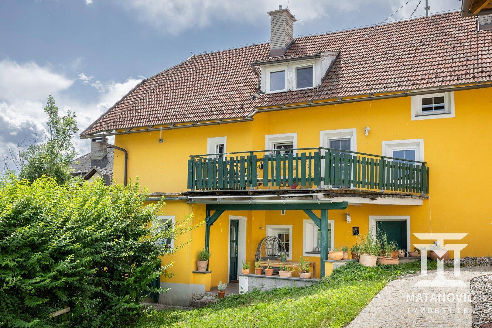 Traditionelles gelbes Haus mit rotem Dach, Balkon und einer überdachten Terrasse im Erdgeschoss.