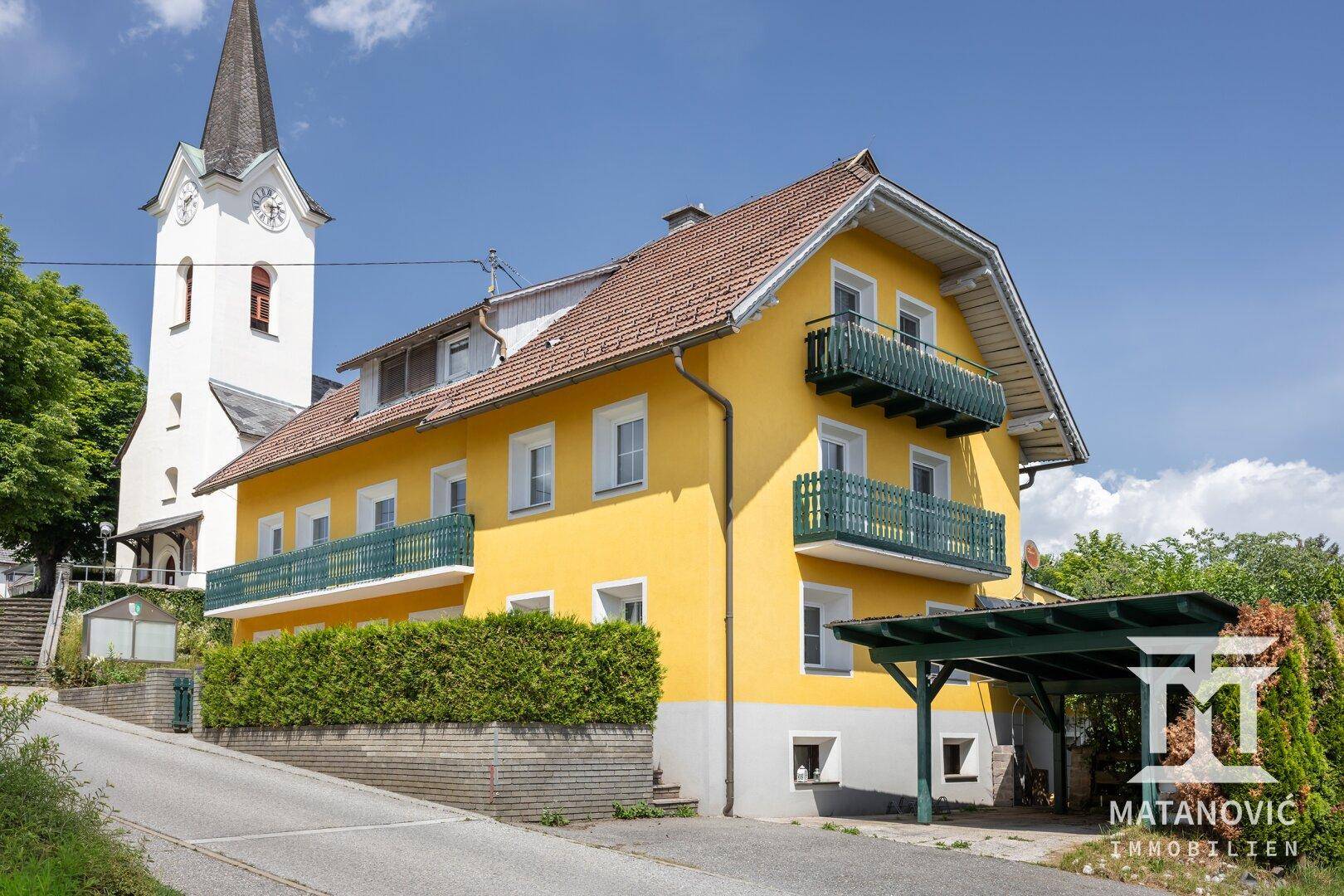Gelbes Haus mit Ziegeldach und Carport, neben einem Kirchturm gelegen, unter blauem Himmel.