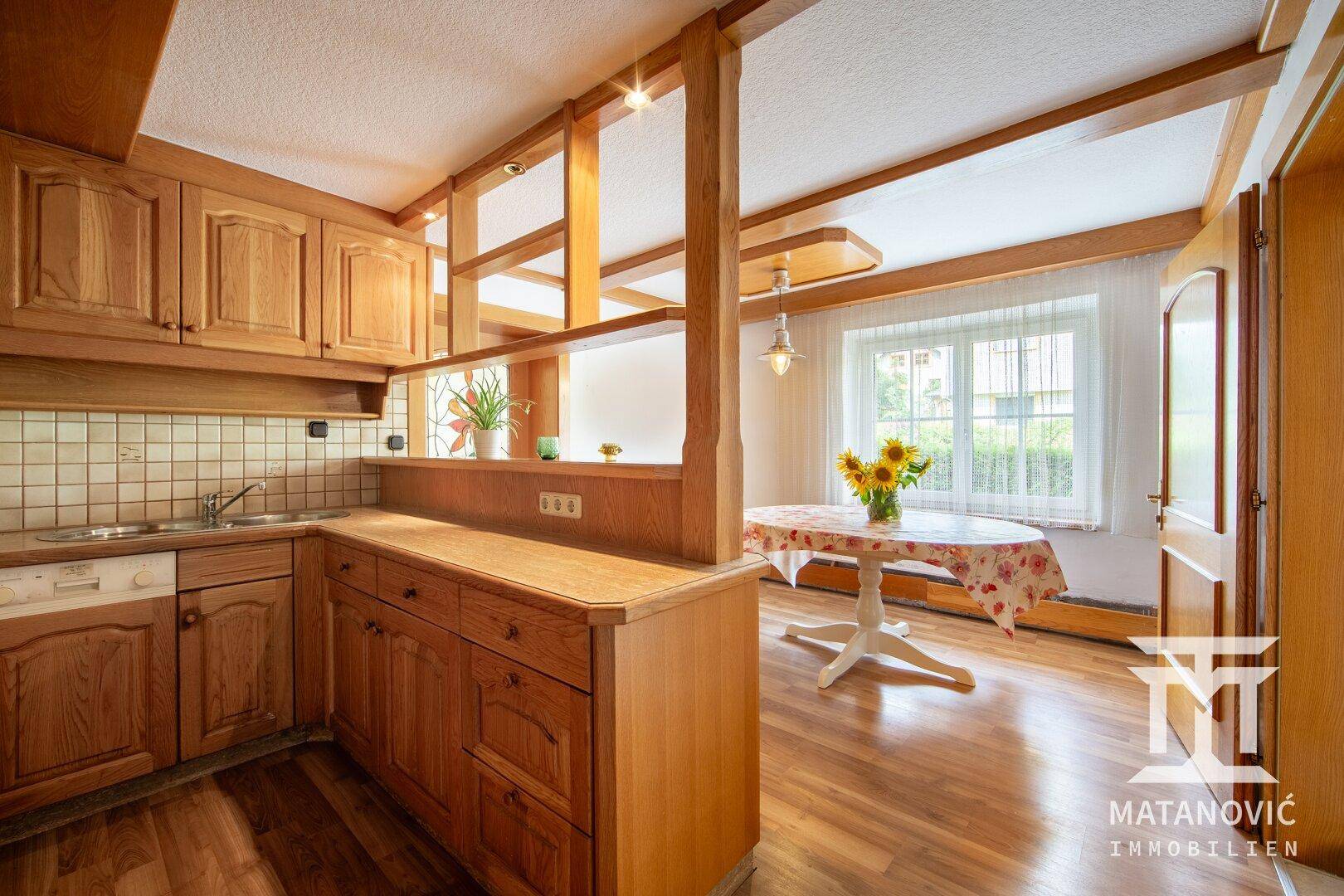 Traditionelle Holzküche mit Essbereich, Einbauschränken und Blick ins Grüne durch das Fenster.