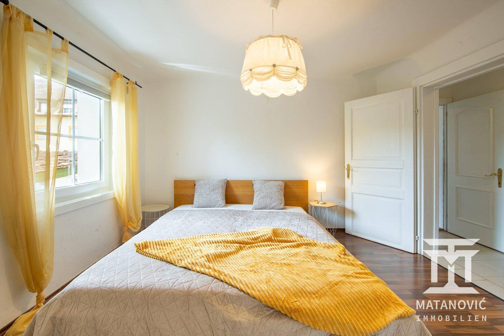 Helles Schlafzimmer mit Doppelbett, Laminatboden und einem Fenster mit gelben Vorhängen.
