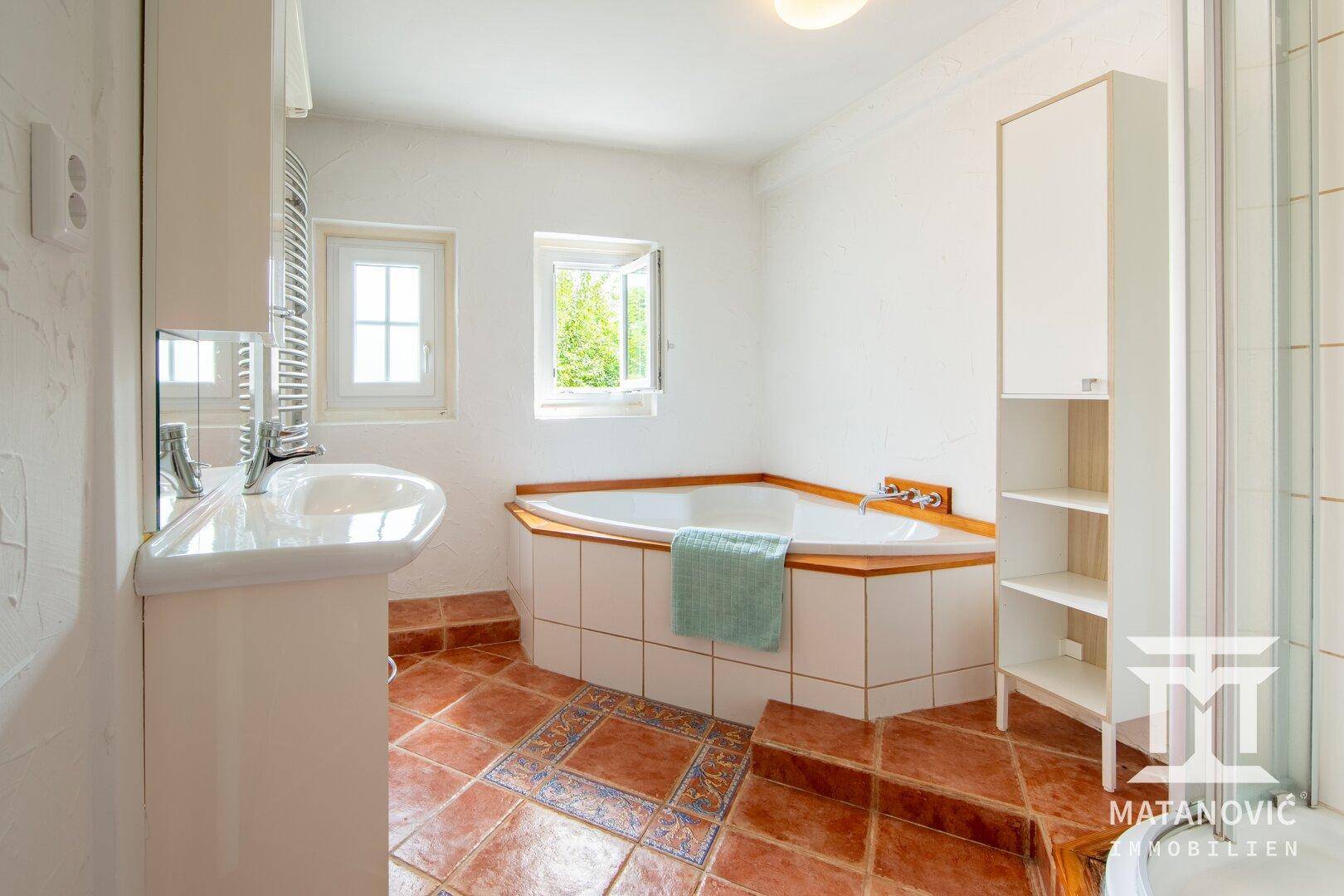 Badezimmer mit Terrakottafliesen, Eckbadewanne, Einzelwaschbecken und zwei Fenstern.