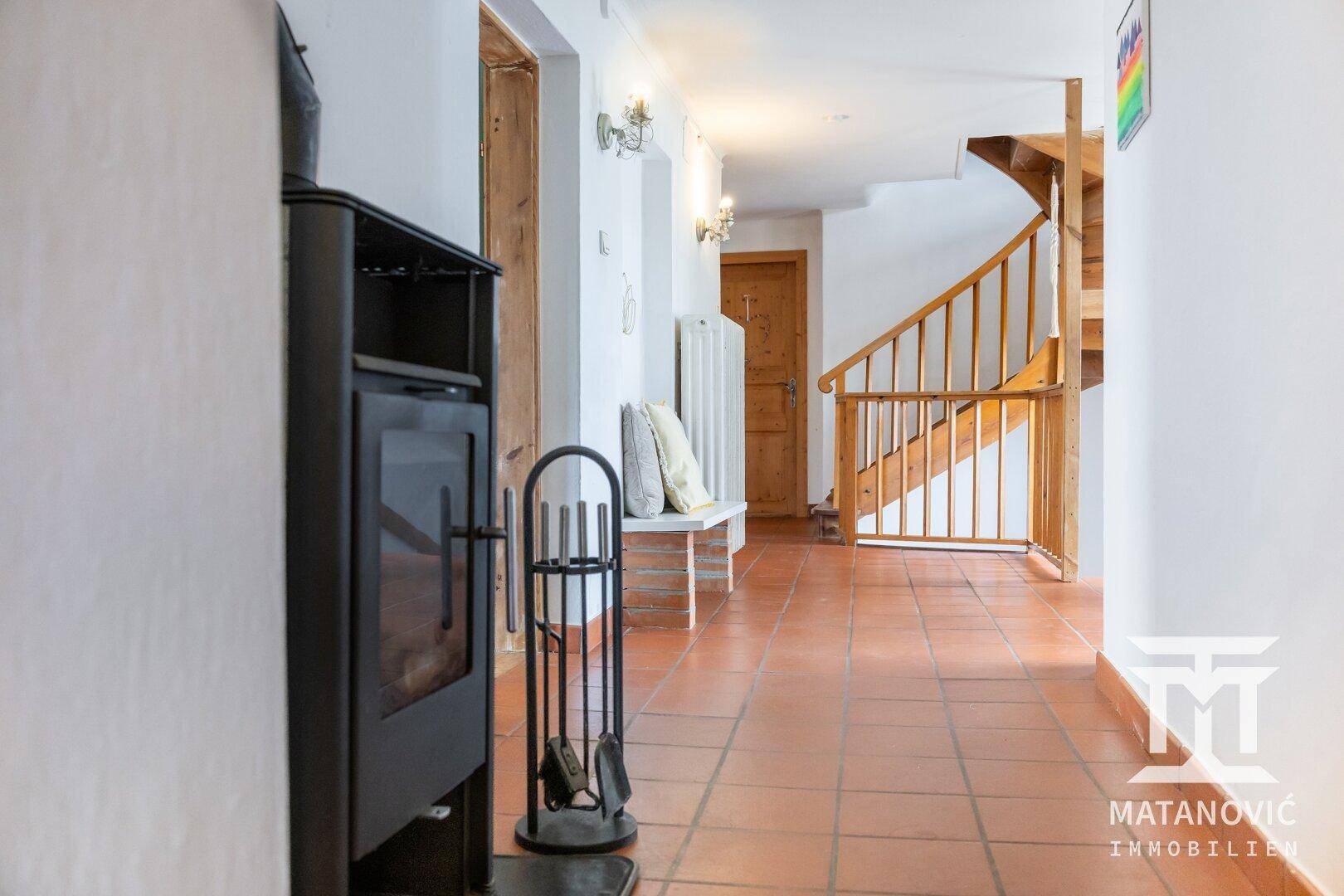 Geräumiger Flur mit Terrakottafliesen, einem Holzofen und einer Holztreppe im Hintergrund.