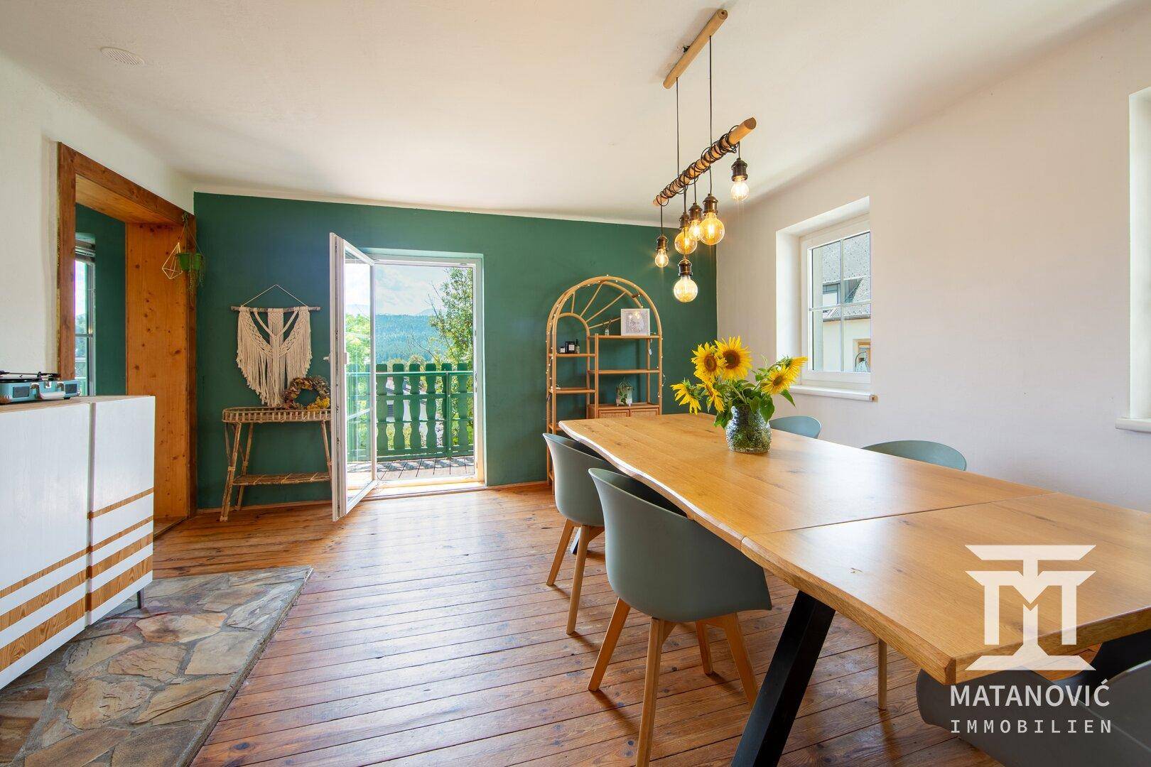 Esszimmer mit großem Holztisch, Dielenboden, grüner Akzentwand und Balkonzugang.