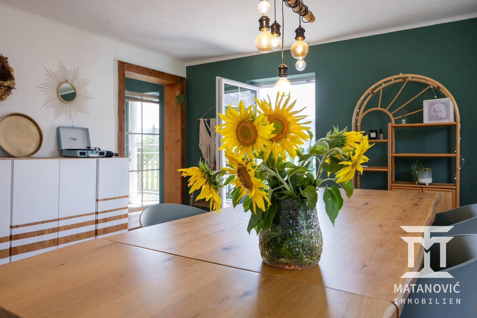 Nahaufnahme des Esstisches mit Sonnenblumen, grüner Wand und Blick auf den Balkon.