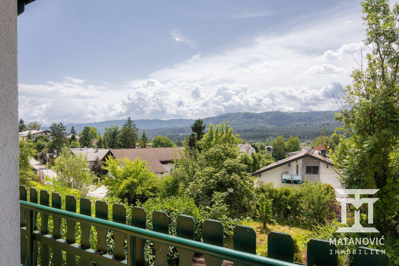 Ausblick vom Balkon auf eine grüne Landschaft mit Häusern und bewaldeten Bergen in der Ferne.