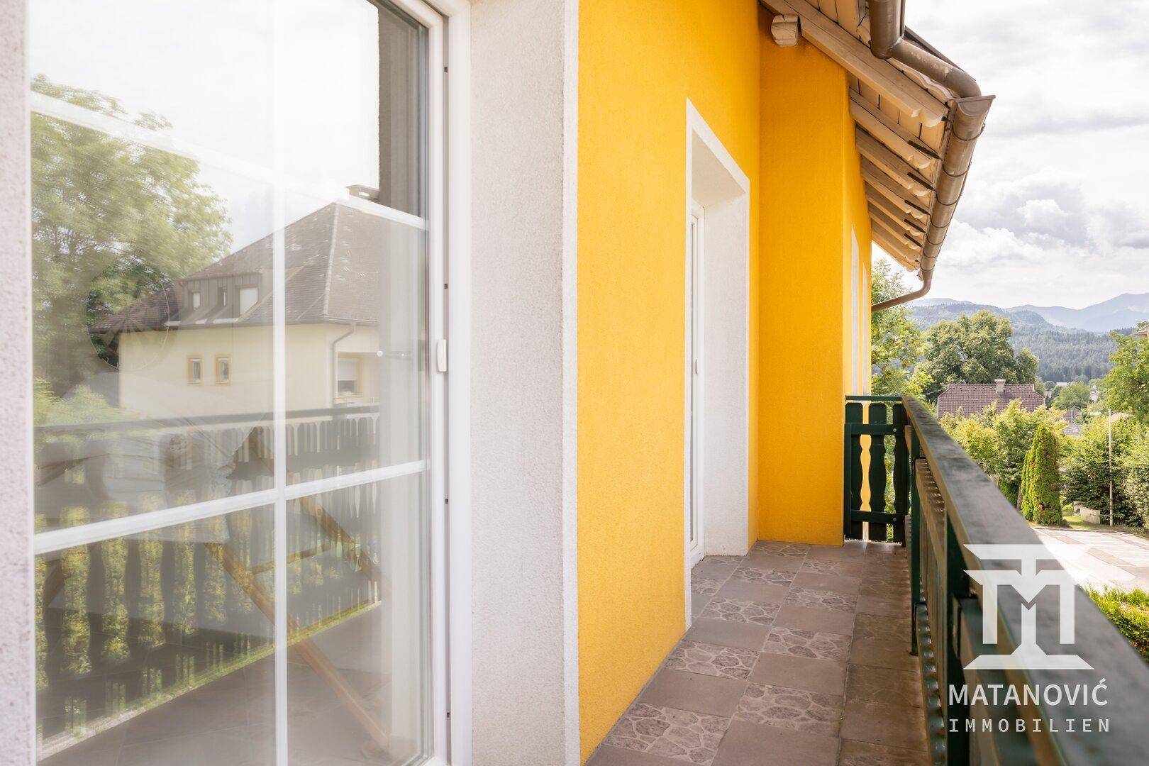 Balkon mit Fliesenboden und grünem Holzgeländer, angrenzend an eine gelbe Hauswand.