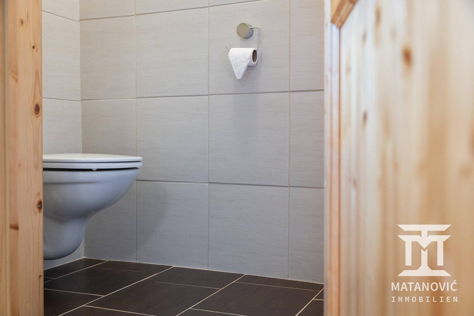 Modernes WC mit wandhängender Toilette, grauen Wandfliesen und dunklem Fliesenboden.