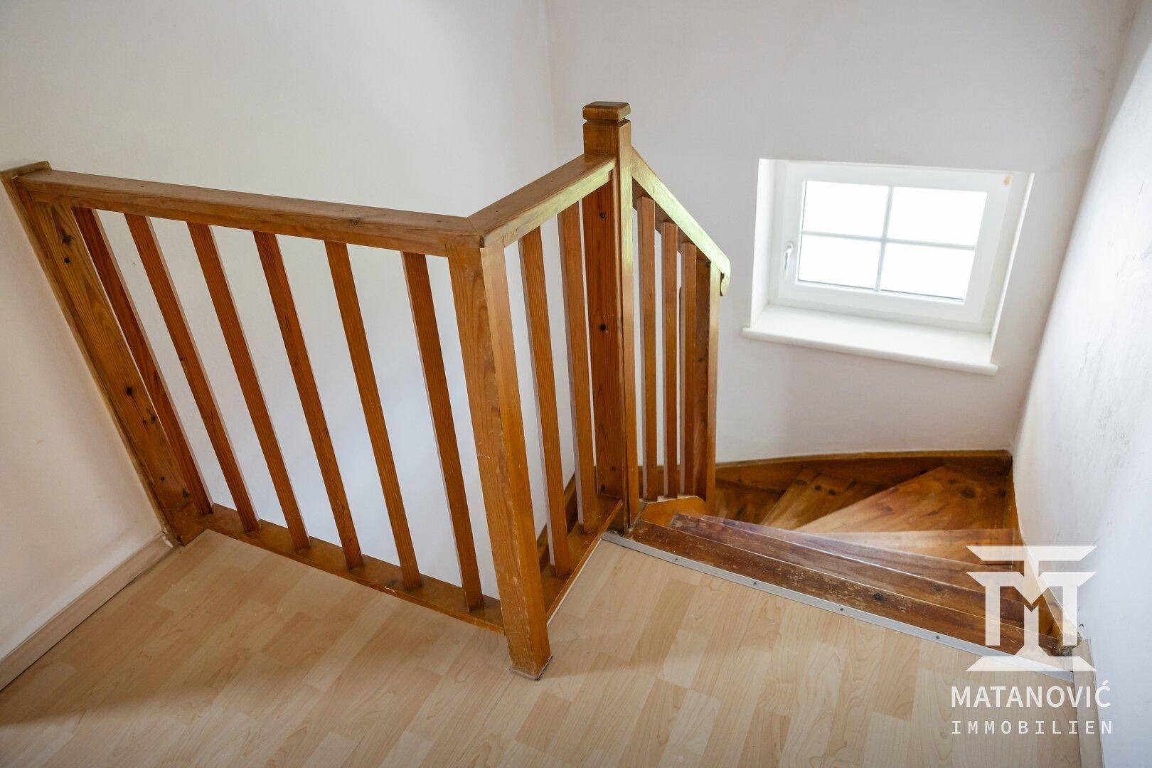 Helle Holztreppe mit Geländer und Fenster, die in ein unteres Stockwerk des Hauses führt.