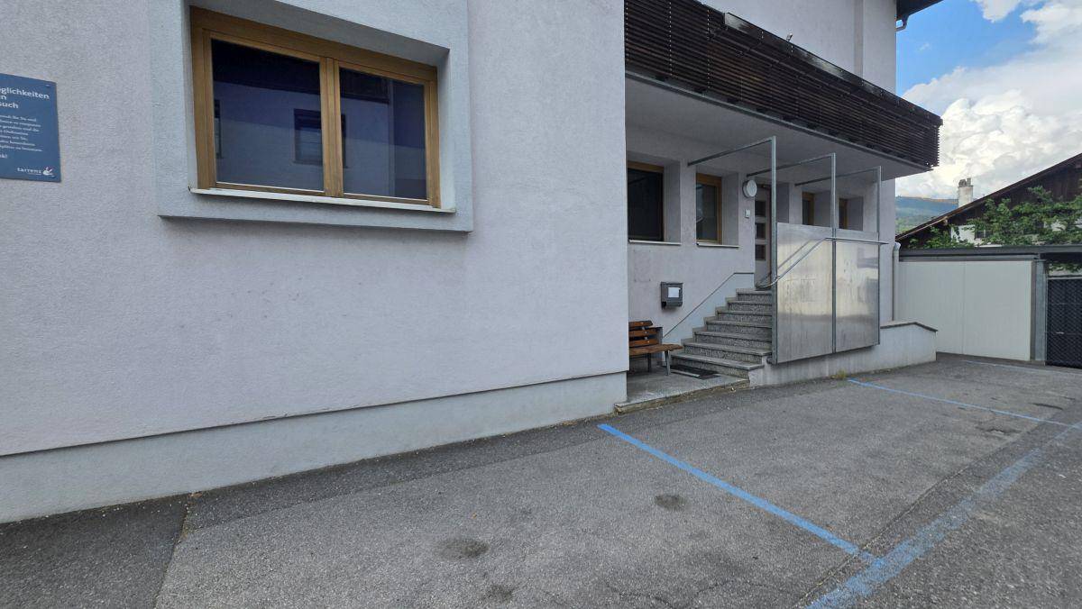 Außenansicht eines Gebäudes mit Eingangsbereich, Treppe und Parkplätzen.