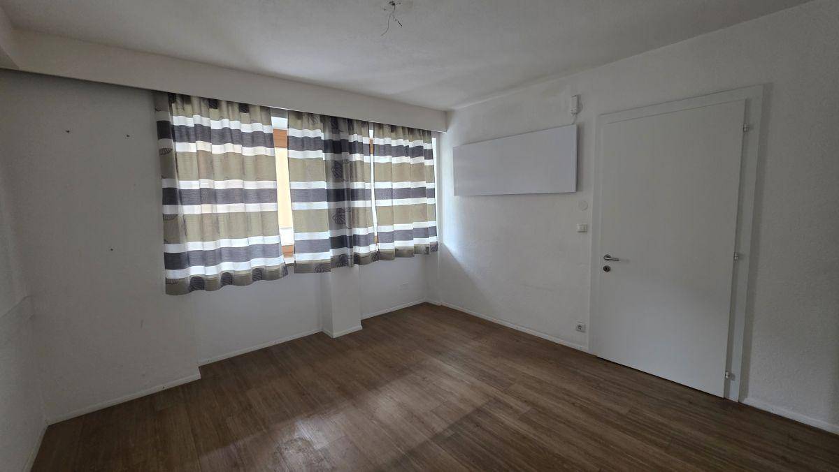 Leerer Raum mit Holzboden, weißen Wänden und einem Fenster mit gestreiften Vorhängen.