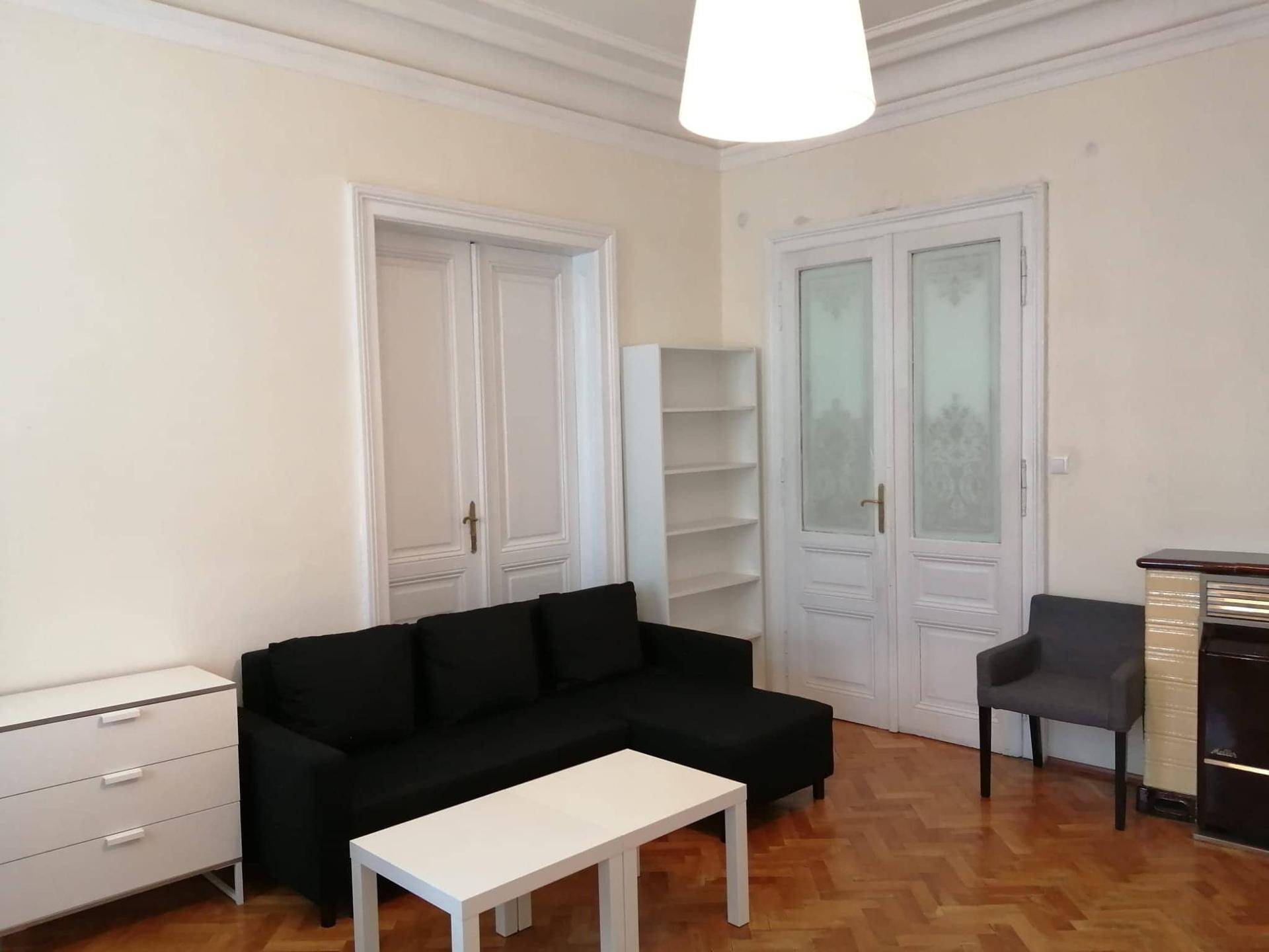 Gemütliches Wohnzimmer mit schwarzem Sofa, Parkettboden und hohen Decken, ergänzt durch einen Couchtisch.