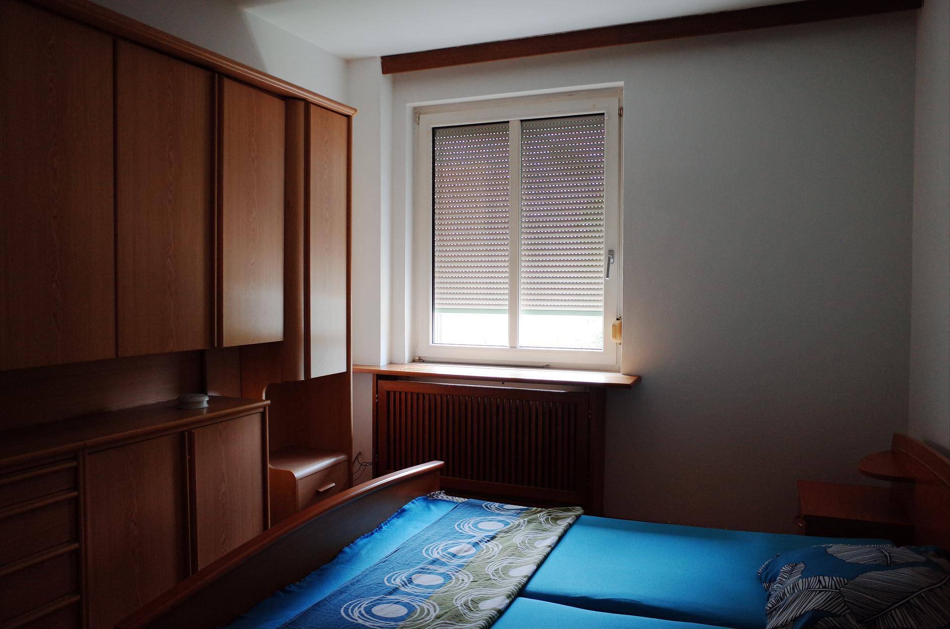Schlafzimmer mit Doppelbett, Einbauschränken aus Holz und einem Fenster mit Rollladen.
