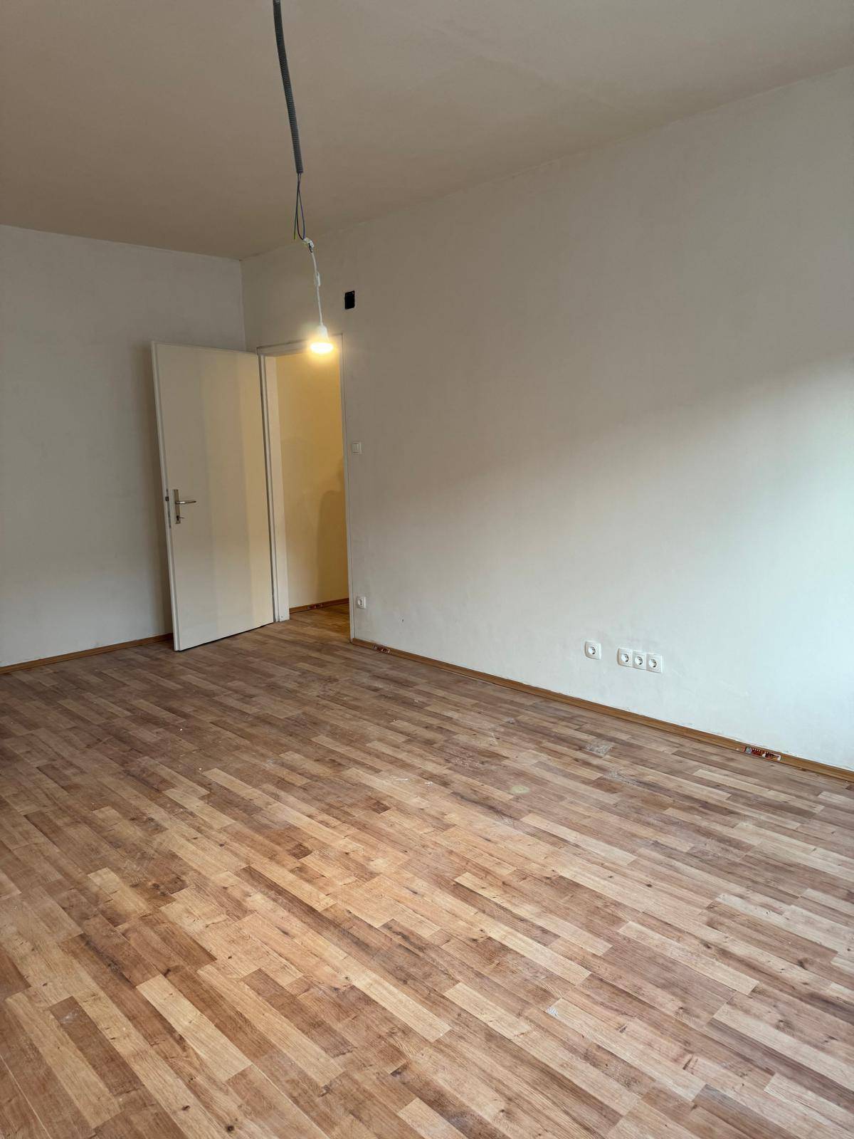 Leerer Raum mit Holzboden und weißen Wänden, eine offene Tür führt in einen weiteren Bereich.
