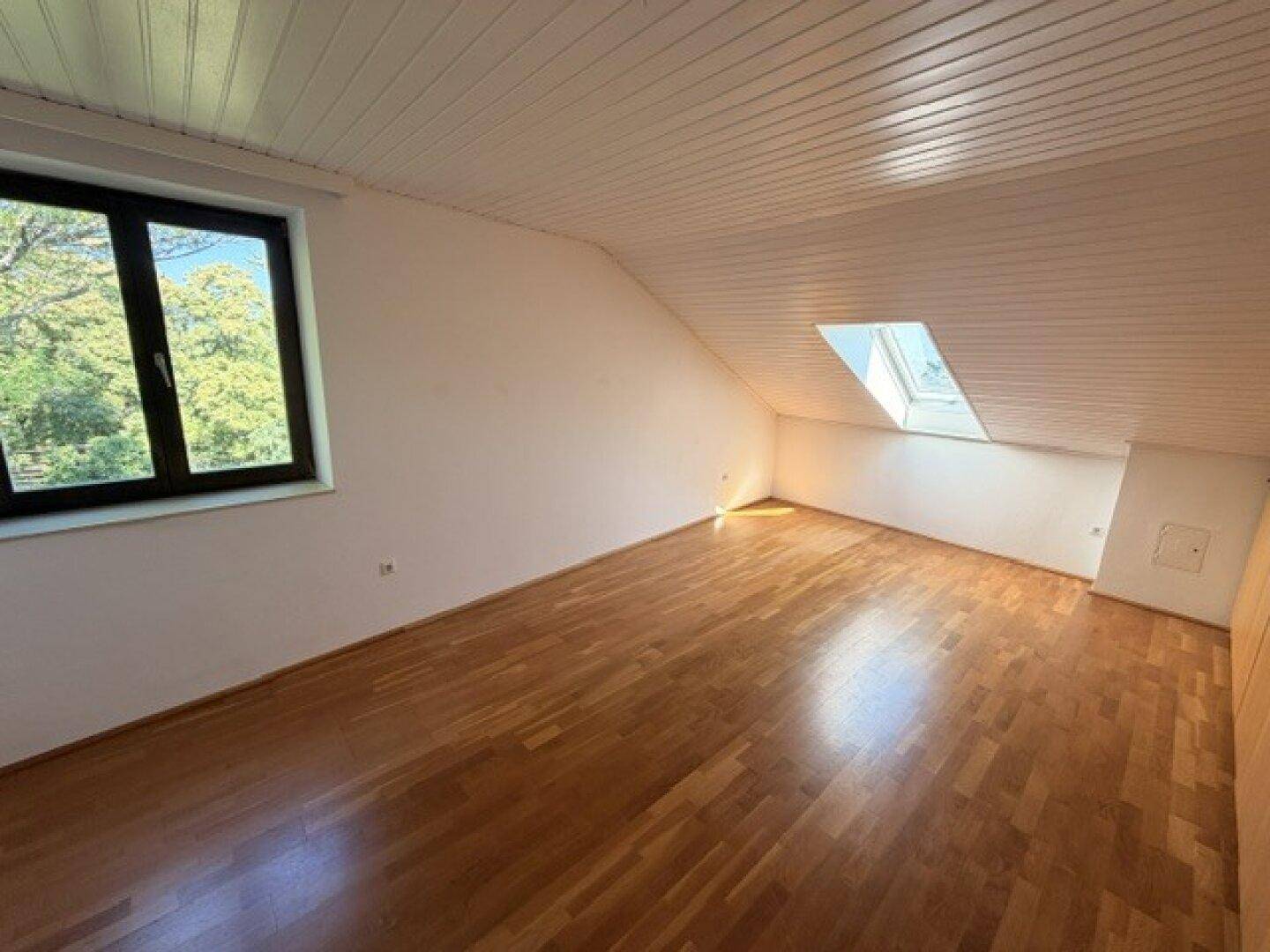 Geräumiger Dachraum mit Holzfußboden, Dachschräge und Blick ins Grüne durch das Fenster.