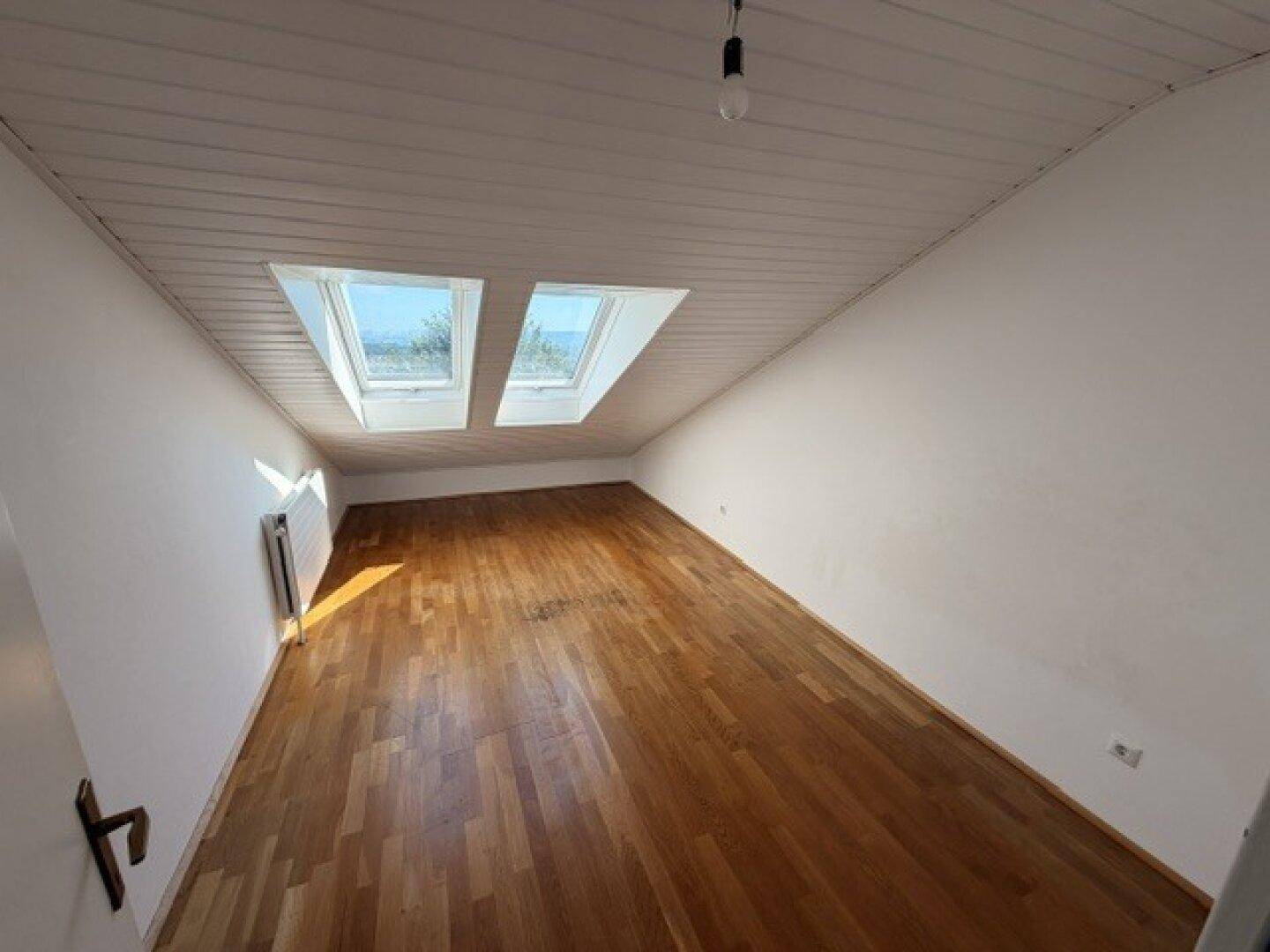 Heller Dachraum mit Holzfußboden, Dachschräge und zwei großen Dachfenstern für viel Licht.